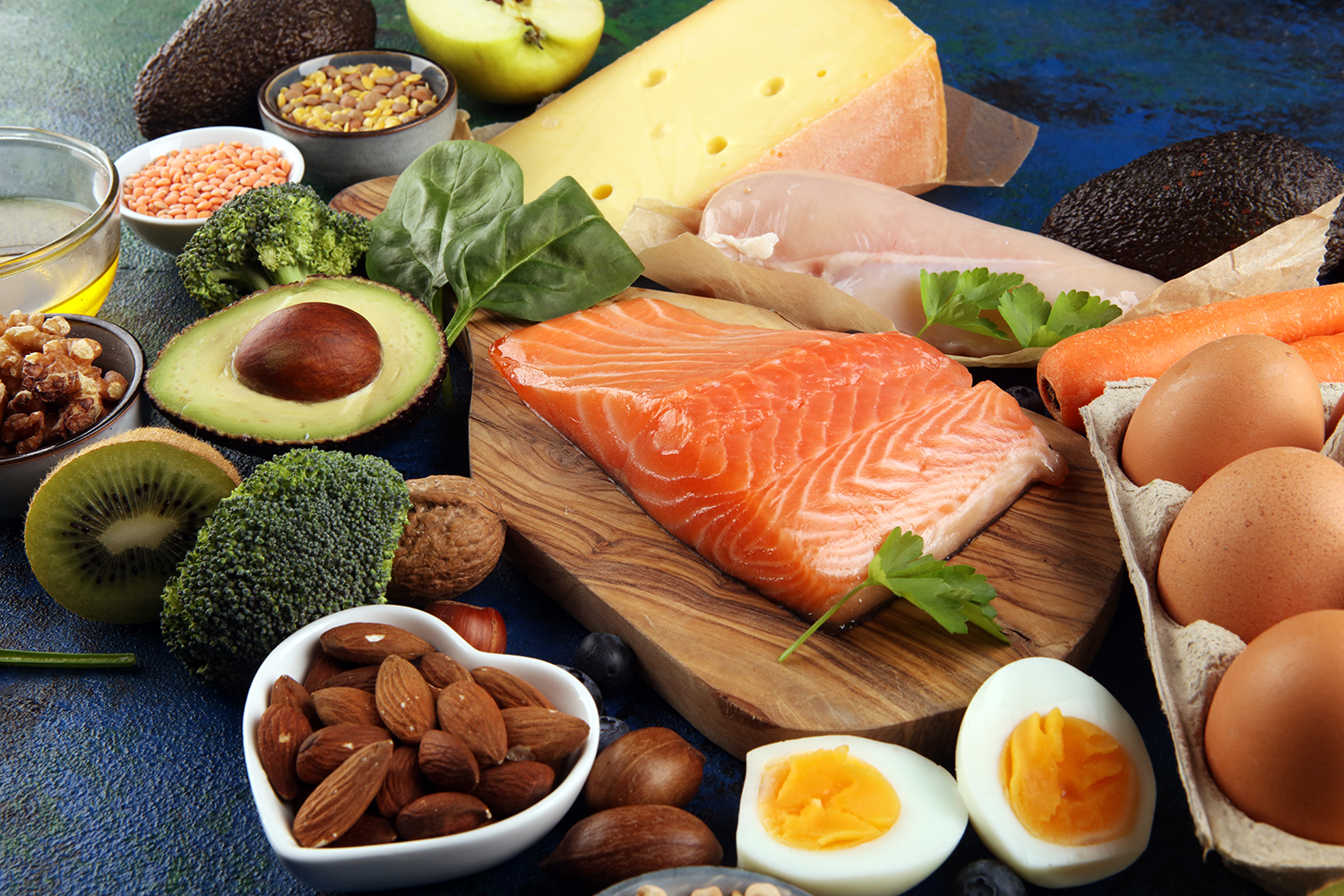 A collection of high protein foods—such as salmon, eggs, almonds, and avocados—are spread out across a table.