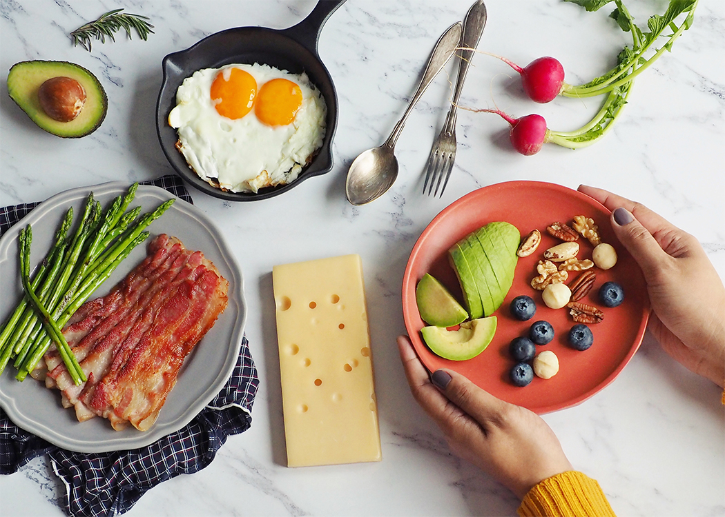 Plates with asparagus, bacon, avocado, nuts, berries, and fried eggs sit on a table that has cheese, half of an avocado, radishes, and silverware on it.