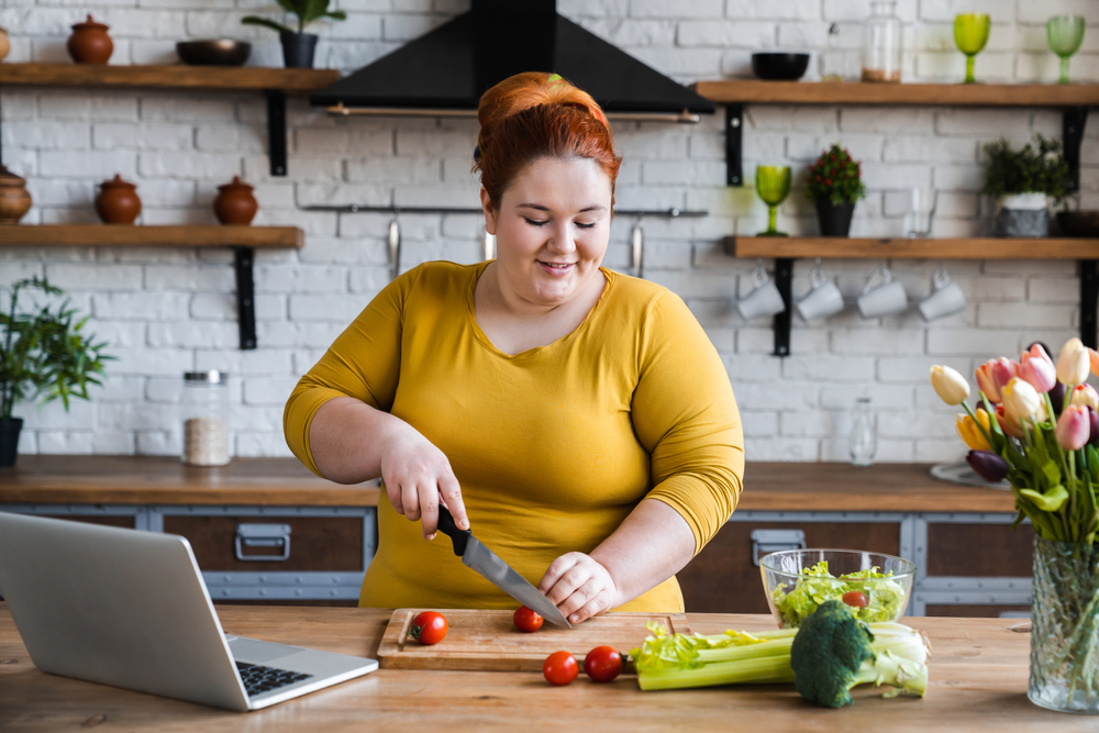 A woman with red hair and a yellow shirt chops vegetables and has her laptop open on the countertop. 