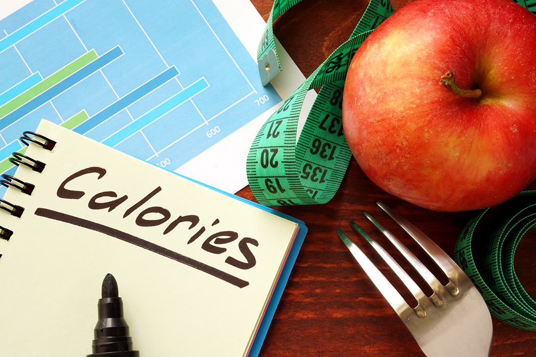A notebook with “Calories” written in it, a chart, a fork, an apple, and a measuring tape are laid out on a table.